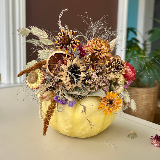 A yellow pumpkin with dried flowers on top of it on a cream table against a brown wall