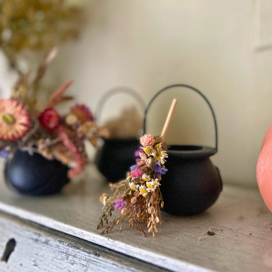 A dried flower broom with a wooden handle and small metal loop, styled on a rustic wooden shelf next to small black cauldrons and dried floral arrangements.