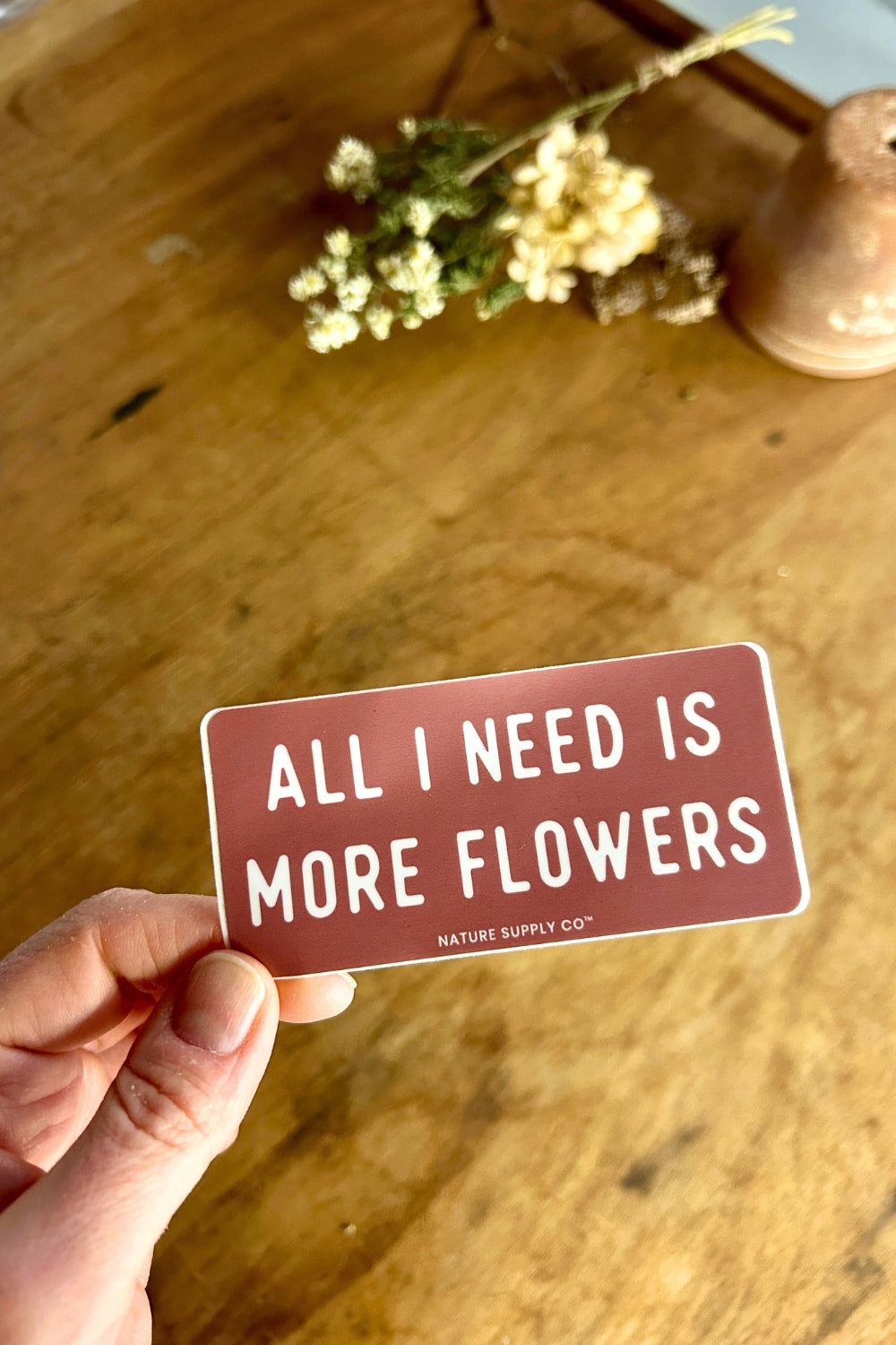Hand holding a sign that says 'All I Need Is More Flowers' on a wooden surface with flowers.